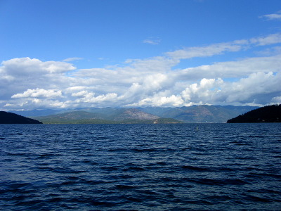 [Looking across a large and what appears to be a very deep body of water. There are mountains on the far side with lots of white clouds near the tops of the mountains but not necessarily all over the sky.]